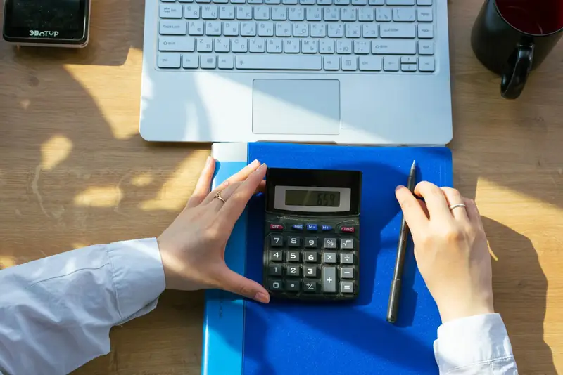 Hands using a calculator beside a laptop and planning folder